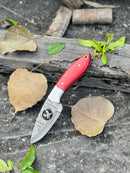 The Texan Knives FIXED BLADE POCKET KNIFE, a personalized Damascus steel hunting knife with a red handle, rests on a weathered log among dried and green leaves on a dark, textured surface.