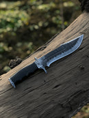 The Texan Knives Best Bowie Knife – a 13" handmade Damascus steel blade with a resin horn handle and leather sheath – rests on a weathered log outdoors, with blurred greenery in the background.