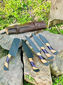 Texan Knives’ Best Knife Block Set features 13" forged stainless steel chef knives with olivewood and epoxy resin handles, displayed outdoors with a leather sheath, surrounded by rocks, grass, and stones.