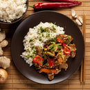 A dark plate with white rice and stir-fried beef featuring red peppers and herbs, delicately enhanced by The Spice Lab Organic Ground Ginger, is surrounded by a bowl of rice, fresh red chilies, garlic, an onion, chopsticks on a bamboo mat, and a jar of The Spice Lab’s signature ginger spice.