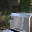 A Myron Mixon BARQ 2400 Pellet Smoker (BARQ-2400) with Wi Fi by Myron Mixon Smokers sits outdoors in a garden with green trees, grass, and a wooden fence in the background, partially in sunlight.