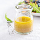 A small glass jar of The Spice Lab Diced Orange Peel sits on a white surface next to a metal whisk, with a plate of salad blurred in the background, hinting at its vibrant citrus aroma.
