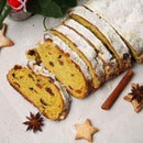 A sliced loaf of fruitcake dusted with powdered sugar is displayed with dried fruit pieces inside. Surrounding the bread on a light surface are cinnamon sticks, star-shaped cookies, and The Spice Lab Ground Star Anise by The Spice Lab, enhancing the festive scene with aromatic spice and a subtle licorice-like flavor.