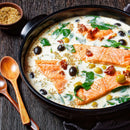 Creamy soup with salmon fillets, black and green olives, sun-dried tomatoes, spinach, and herbs—including a dash of The Spice Lab Ground Oregano—in a black bowl on a wooden table, accompanied by wooden spoons and a small bowl of seasoning nearby.