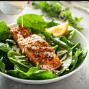 Grilled salmon on a bed of fresh spinach leaves, lightly seasoned with The Spice Lab Ground Thyme from The Spice Lab and drizzled with creamy dressing, with a lemon wedge on the side, served in a white bowl.