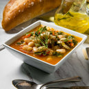 A square bowl of bean and vegetable soup, garnished with herbs and highlighting the mild licorice flavor of The Spice Lab Organic Fennel Seeds, sits next to a loaf of bread, a glass jug of oil, and cutlery on a marble surface.