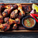 Grilled chicken drumsticks on a wooden tray, seasoned with The Spice Lab Organic Ground Allspice, served alongside a bowl of sauce, two red chili peppers, garlic cloves, and lemon wedges on the side.