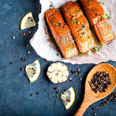 Three cooked salmon fillets on parchment paper, garnished with herbs and a sprinkle of freshly ground pepper from The Spice Lab Premium Kings Peppercorn Grinder, sit next to lemon slices, a halved garlic bulb, scattered mixed peppercorns from The Spice Lab blend, and a wooden spoon filled with colorful peppercorns—all arranged on a dark blue surface.