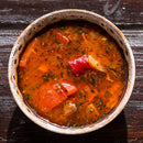A bowl of red soup with chunks of vegetables and herbs, infused with The Spice Lab Organic Spanish Sweet Paprika, sits on a wooden surface.