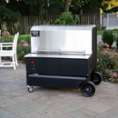 A Myron Mixon BARQ 3600 Pellet Smoker (BARQ-3600) by Myron Mixon Smokers sits on a stone patio with wheels, surrounded by greenery, pink flowers, and a white bench in a garden setting.