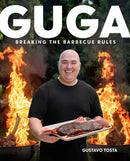 A smiling man in a black shirt holds a tray of grilled meat, standing in front of two flaming grills. Large text reads "Guga: Breaking the Barbecue Rules - Hardcover" by Books by splitShops at the bottom, inspired by Gustavo Tosta’s popular Guga Foods channel and creative barbecue recipes.