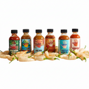 Six vibrant bottles from the Culture Hot Sauce Gift Sampler pack by Culture Hot Sauce are lined up in a row behind a mound of fresh light green chili peppers, displayed against a clean white background—the perfect sampler set for hot sauce enthusiasts.