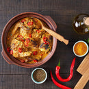 A clay pot brimming with curry chicken, garnished with vibrant red chili slices and a touch of The Spice Lab Ground Coriander, surrounded by an array of spices, whole red chilies, and a bottle of oil on a dark wooden table—a bold dish that’s perfect for BBQ restaurants looking to add flavor with The Spice Lab Ground Coriander 1 lb bulk foodservice spice.
