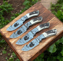 Three Texan Knives Custom Handmade Guthook Hunter Knives by Texan Knives, featuring Damascus steel blades and stag horn handles, are displayed side by side on a wooden board outdoors amid lush green foliage.