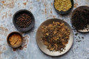 World Spice Asian Tea Rub by World Spice features a vibrant blend of coriander seeds, peppercorns, dried herbs, and a touch of Lapsang Souchong, artfully arranged in bowls and plates on a textured surface—the perfect combination for adding a subtle smoky flavor to your next unique tea rub.