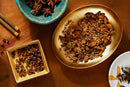 An overhead view of spices on a wooden table, featuring star anise in a blue bowl, World Spice Chinese Five Spice by World Spice on a gold plate, aromatic Sichuan peppercorns in a square dish perfect for enhancing any stir-fry, and additional spices arranged in a white dish.