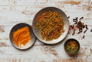 Three bowls rest on a rustic white surface: one filled with vibrant orange turmeric powder, another containing World Spice Khmeli Suneli—a traditional Georgian spice blend from World Spice featuring a medley of colorful dried spices—and a small bowl of fresh green herbs. Whole cloves are scattered beside the bowls.