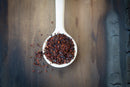 A white spoon filled with crushed red chili flakes rests on a rustic wooden surface, with some of the vibrant World Spice Limón-Chile blend by World Spice scattered beside it.