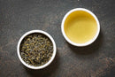 A bowl of loose World Spice Nepalese Green tea leaves from World Spice rests beside a cup of freshly brewed green tea on a dark, textured surface.
