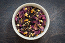 A bowl filled with a vibrant loose leaf tea blend—featuring dried rose petals, orange peels, spices, and herbs—rests on a dark, textured surface. The World Spice Loose-Leaf Tea Tasting Flight by World Spice makes an ideal addition to any herbal tea sampler or a thoughtful tea gift set.
