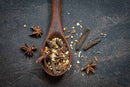 A wooden spoon filled with crushed spices, including star anise and long pepper, rests on a dark surface alongside scattered whole spices, capturing the aromatic base of World Spice Pho-Nominal Broth by World Spice.
