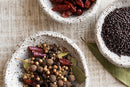 Three stone bowls hold whole spices: one with dried chilies, one with black mustard seeds, and one with World Spice Pickling Spice from World Spice—a vibrant blend of coriander seeds, allspice, peppercorns, bay leaves, and dried chilies. Beautifully arranged and viewed from above, this enticing array evokes the fragrant condiments used for traditional British classics.