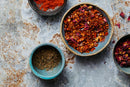 Three ceramic bowls with spices: one containing World Spice Rose Harissa by World Spice, a vibrant citrus and floral blend of dried chili flakes and rose petals; one with cumin seeds; and one Ottolenghi-inspired bowl filled with bright ground red spice, all displayed on a rustic gray surface.