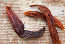 Three dried World Spice Aji Mirasol chiles from World Spice rest on a rustic, faded light brown wooden surface with visible texture and grain, highlighting their distinctive fruity flavor and mild chile appeal.