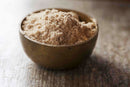 A brass bowl filled with light brown, finely ground World Spice Amchoor from World Spice sits on a rustic wooden surface in soft light—a staple ingredient that adds tangy depth to traditional Indian cuisine.