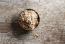 A round metal container of World Spice Amchoor by World Spice rests on a rustic wooden surface, with a small amount of the light brown, tangy unripe mango powder spilled nearby.