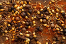 A close-up of assorted aromatic spices—including coriander seeds, black peppercorns, cloves, allspice, and cumin seeds—classic ingredients in Middle Eastern spice blends like World Spice Baharat by World Spice—scattered on a wooden surface dusted with ground spices.