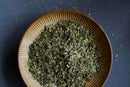 A brown ceramic bowl filled with World Spice Basil by World Spice, perfect for flavoring Mediterranean dishes, sits on a gray surface, with some of the dried green basil scattered around the bowl.