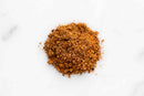 A small pile of World Spice Fin & Feather BBQ Rub from World Spice, with its finely ground brown seasoning, is placed on a white surface.