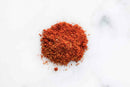 A small pile of World Spice Smoky BBQ Rub by World Spice, a finely ground red-orange spice mix, is spread on a white marble surface.