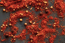 Ground red spice with whole coriander and peppercorns scattered on a dark surface, highlighted by the bold flavors of World Spice Smoky BBQ Rub from World Spice for an irresistible smoky barbecue effect.