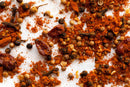 A close-up of World Spice Berbere by World Spice—a traditional Ethiopian blend featuring ground red chili powder, coriander seeds, whole cloves, black peppercorns, and dried red chilies—scattered on a white surface.