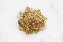 A small pile of World Spice Biscayne Citrus Rub from World Spice, featuring dried Italian seasoning herbs and spices with visible green herb flakes and red pepper bits, offers a zesty twist as it rests on a white marble surface.