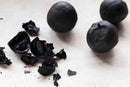 Three whole World Spice Black Lemons from World Spice, along with several broken pieces, are displayed on a light-colored surface. Their rough, dark exteriors and fragmented interiors are shown—perfect as a traditional souring agent in Middle Eastern cuisine.