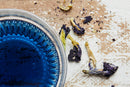 A close-up shows a glass bowl filled with vibrant blue liquid from World Spice Butterfly Pea Flower, a striking cocktail ingredient also called blue tea, positioned on the left. On the right, dried World Spice Butterfly Pea Flowers are scattered across a rustic, distressed wooden surface.