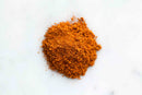 A small mound of World Spice Caribbean Spice, an orange-brown powder from World Spice reminiscent of aromatic Caribbean seasoning, sits on a white surface.