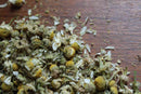 Loose World Spice Chamomile flowers from World Spice are scattered on a wooden surface, their yellow centers and white petals embodying the calming essence of this classic relaxing tea.