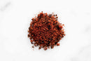 A small mound of World Spice New Mexico Chili Powder, a reddish-brown ground chili spice by World Spice, is spread on a white surface. The powder has a coarse texture with visible flecks of darker and lighter pieces throughout—perfect for adding depth of flavor to tacos or fajitas.
