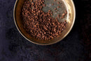 A brass bowl filled with cacao nibs sits on a dark, textured surface. The small, brown, and irregularly shaped cacao nibs partially cover the ornate pattern inside the bowl. Nearby, World Spice Cinnamon Cassia Chips from World Spice are scattered, hinting at a blend of flavors reminiscent of chai masalas and mulling spices.