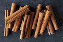 A bundle of World Spice Cinnamon Stick by World Spice rests on a dark, textured surface. These brown, cylindrical sticks vary slightly in size and appearance—ideal for adding rich flavor to savory recipes or infusing mulled drinks with their signature warm aroma.