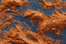 Coarsely ground orange-brown World Spice Elliott Bay Seasoning from World Spice is scattered across a dark blue surface in uneven piles and specks, highlighting the blend’s versatility—perfect for enhancing a wide variety of seafood dishes.