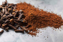 Whole cloves and ground cloves from World Spice are displayed on a gray surface. The World Spice whole cloves are dark brown with lighter tips, while the ground cloves from World Spice form a small, loose pile of fine brown powder, ready to add their intense flavor to any dish.