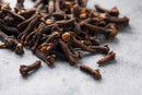 A close-up of World Spice Cloves by World Spice, celebrated for their intense flavor, scattered across a textured gray surface, capturing the rich heritage of the spice islands.