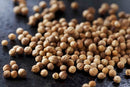 A scattering of World Spice European Coriander Seed by World Spice lies on a dark surface. The round, light brown seeds display their characteristic textured appearance, with some grouped together and others spread loosely.