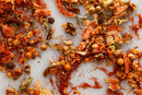 A close-up of assorted dried spices and herbs—including World Spice Creole Spice by World Spice—featuring Creole seasoning, red chili powder, dried onions, green herbs, and whole peppercorns scattered on a white surface. This vibrant blend highlights subtle trinity mix notes and a gentle hint of peppery heat.