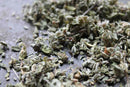 A close-up of World Spice Damiana from World Spice, showing dried, crumbled green herbs scattered on a gray surface, suggesting their use in herbal beverages celebrated for distinctive flavor.
